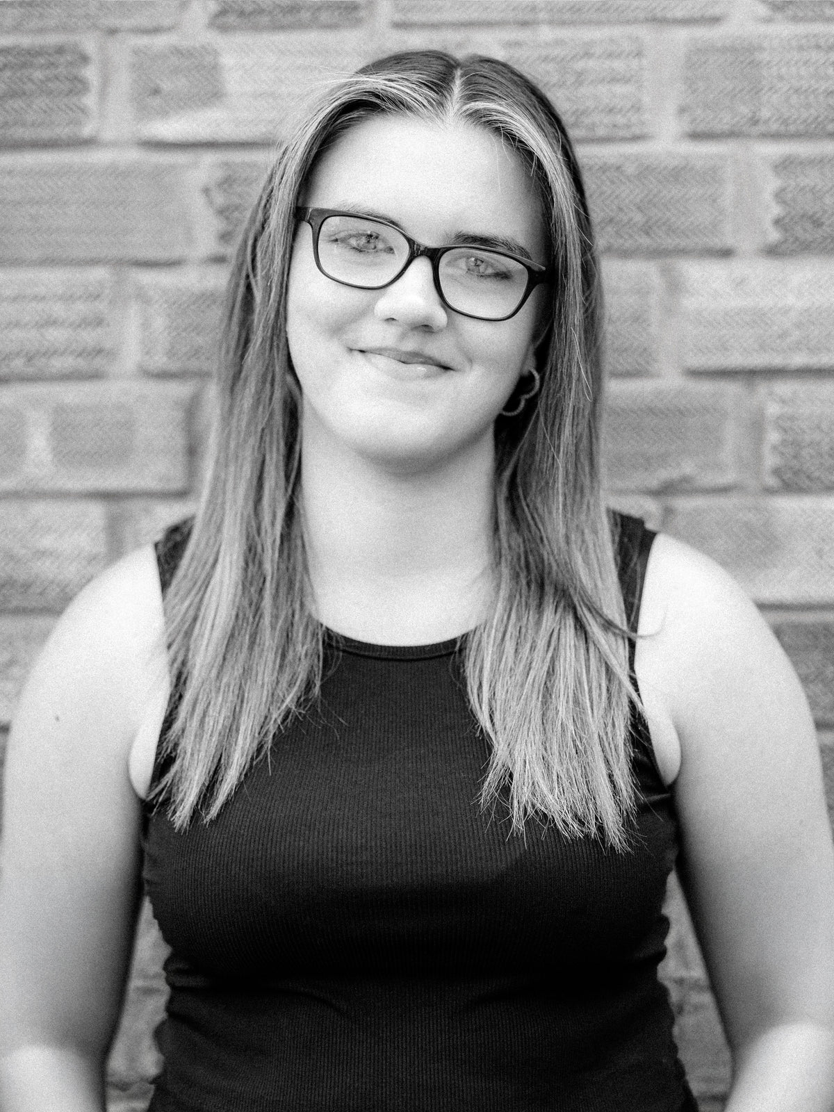 A black and white portrait of Method stylist Abbie, a young woman with long hair and glasses, wearing a dark ribbed tank top and standing against a brick wall background.