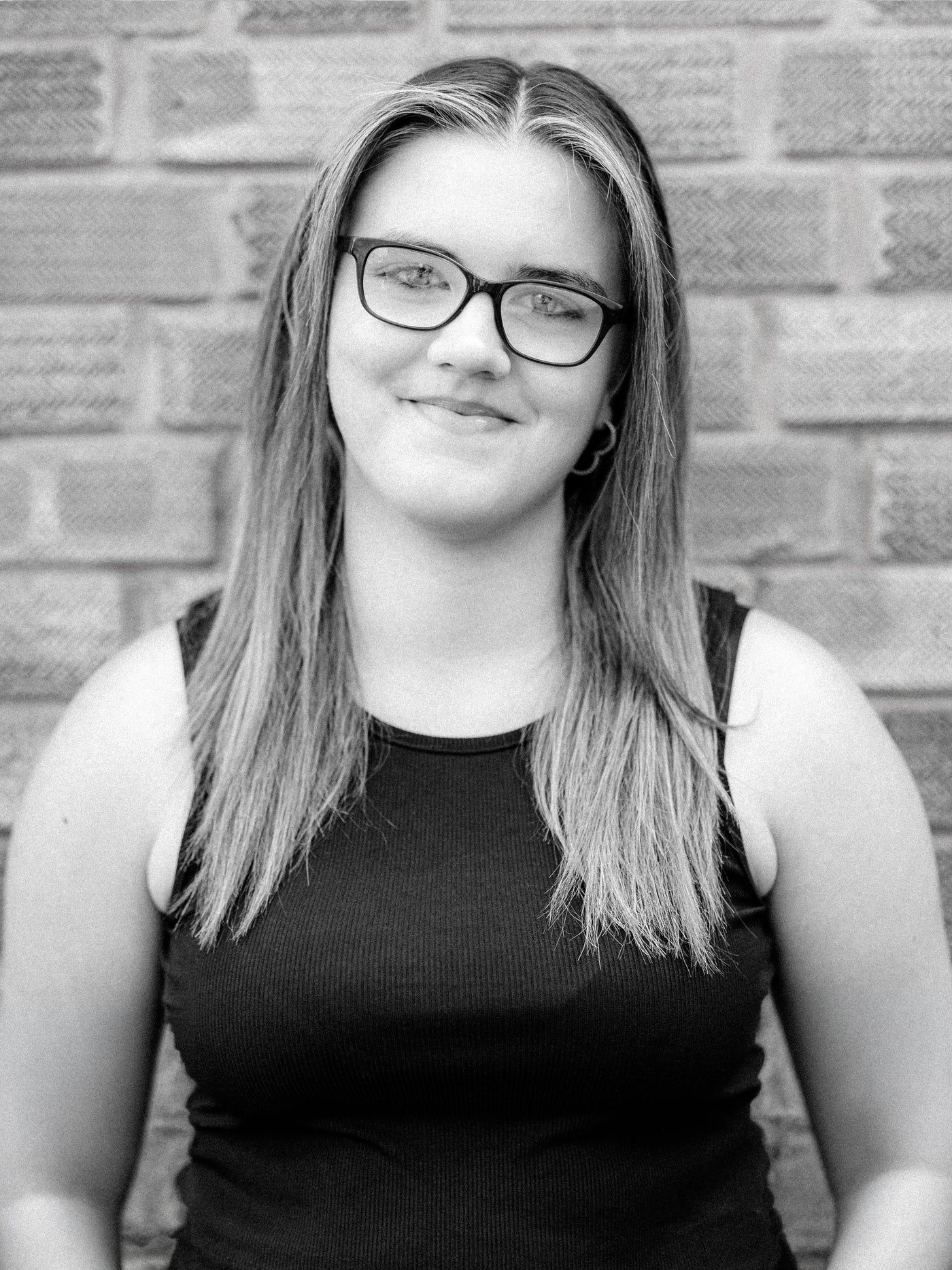 A black and white portrait of Method stylist Abbie, a young woman with long hair and glasses, wearing a dark ribbed tank top and standing against a brick wall background.