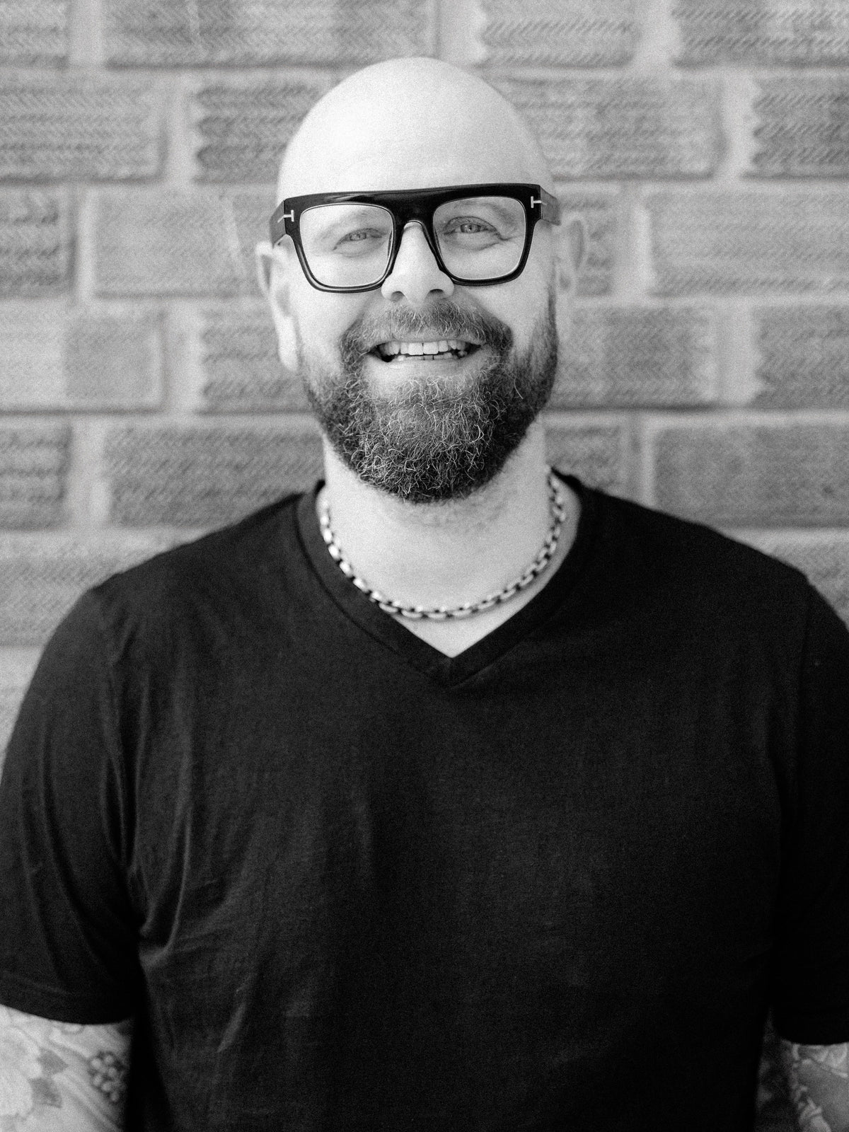 A black and white portrait of Method stylist Daniel, a smiling man with a beard, wearing thick black-rimmed glasses, a silver necklace, and a black v-neck t-shirt, standing in front of a brick wall.