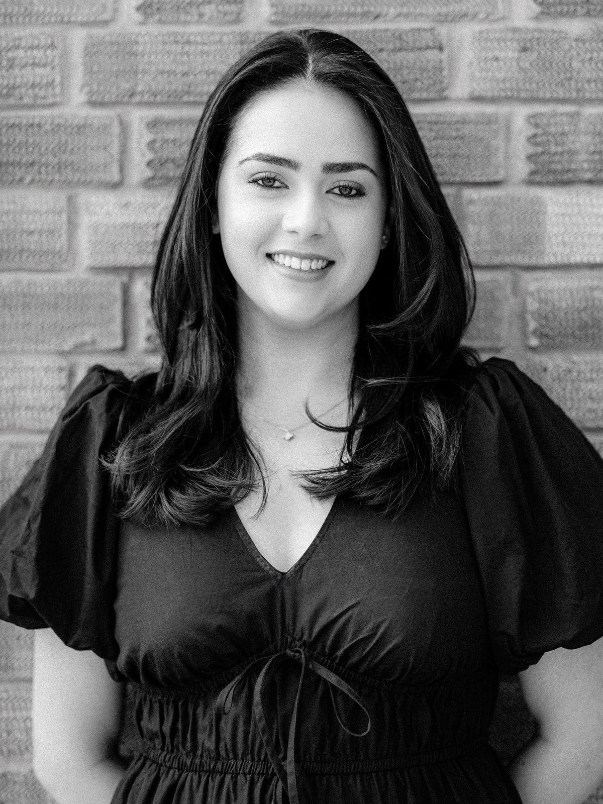 Senior stylist Fizz, a smiling woman with long dark hair stands in front of a brick wall, wearing a black V-neck dress with puffed short sleeves and a drawstring at the waist. The photo is in black and white.