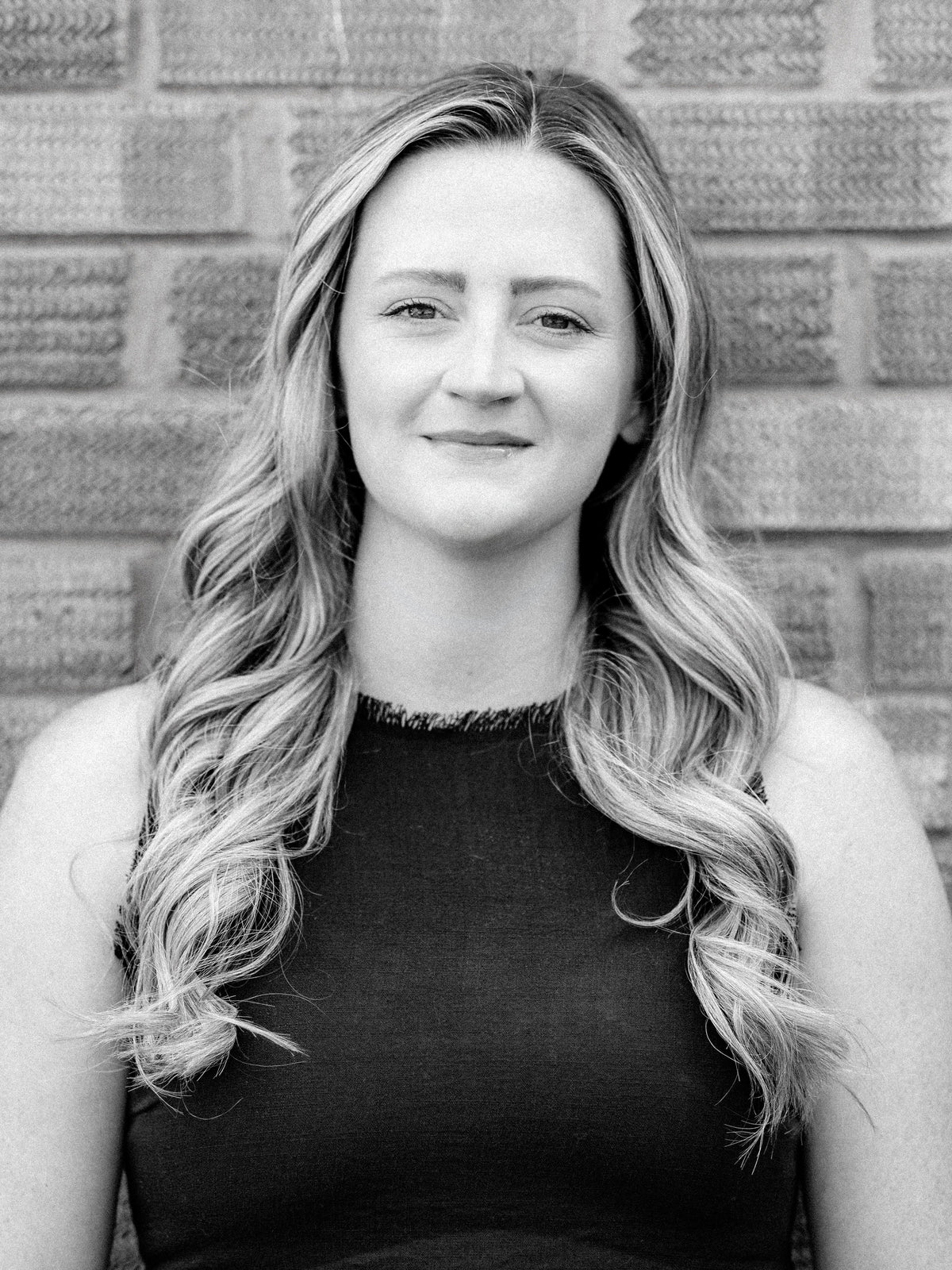 A black and white portrait of senior stylist and colourist Fran, a woman with long wavy hair wearing a sleeveless dark top, standing in front of a brick wall.