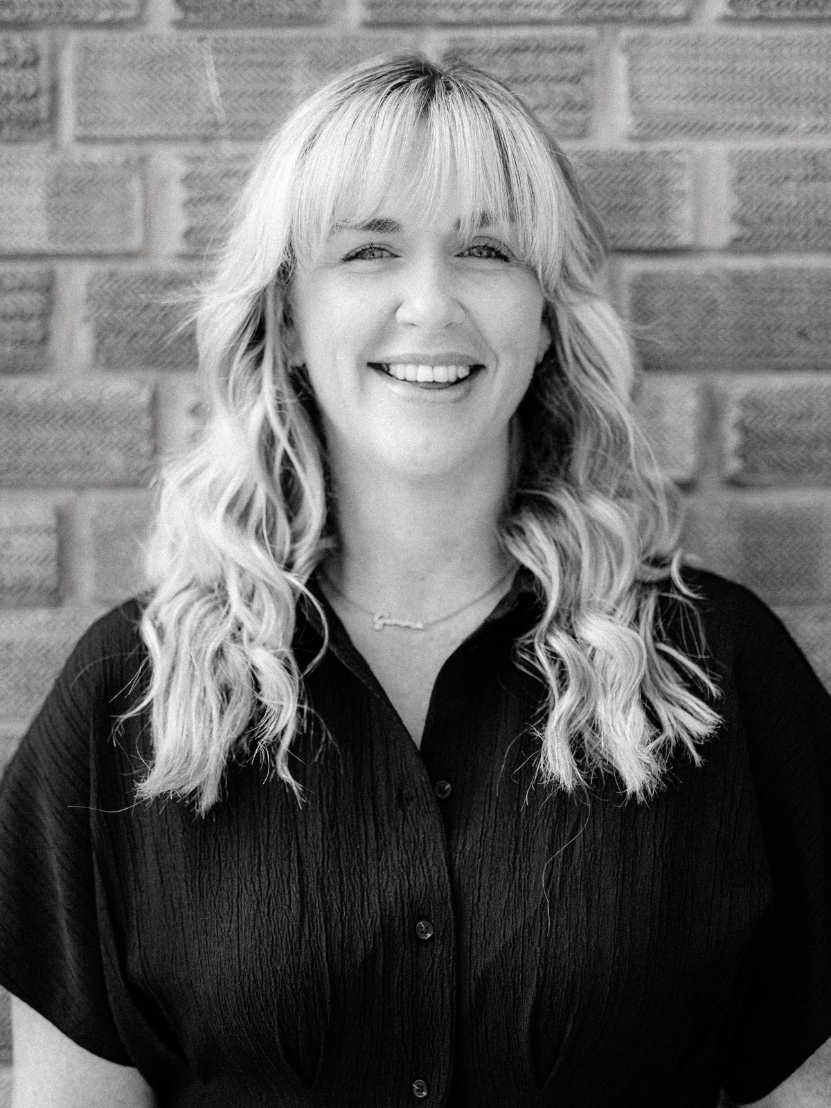 A black and white portrait of Gemma, senior stylist and colourist at Method Hair Salon, a smiling woman with long blonde wavy hair, wearing a black shirt and standing in front of a brick wall.