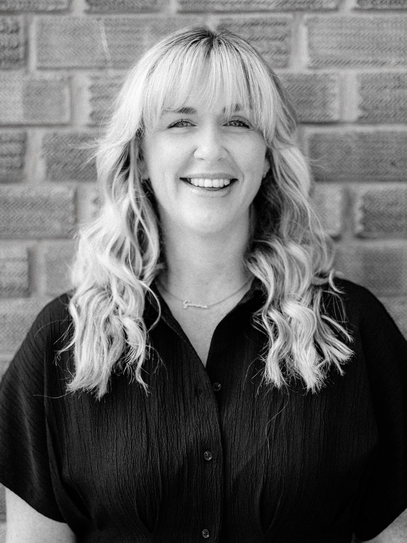A black and white portrait of Gemma, senior stylist and colourist at Method Hair Salon, a smiling woman with long blonde wavy hair, wearing a black shirt and standing in front of a brick wall.