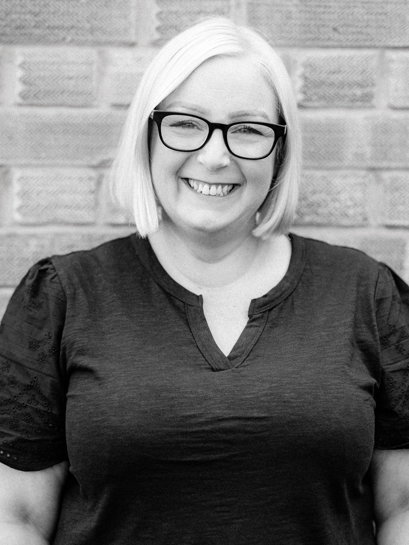 Master colourist Gillian, a smiling woman with short hair and glasses, wearing a black top and standing in front of a brick wall. The image is in black and white.