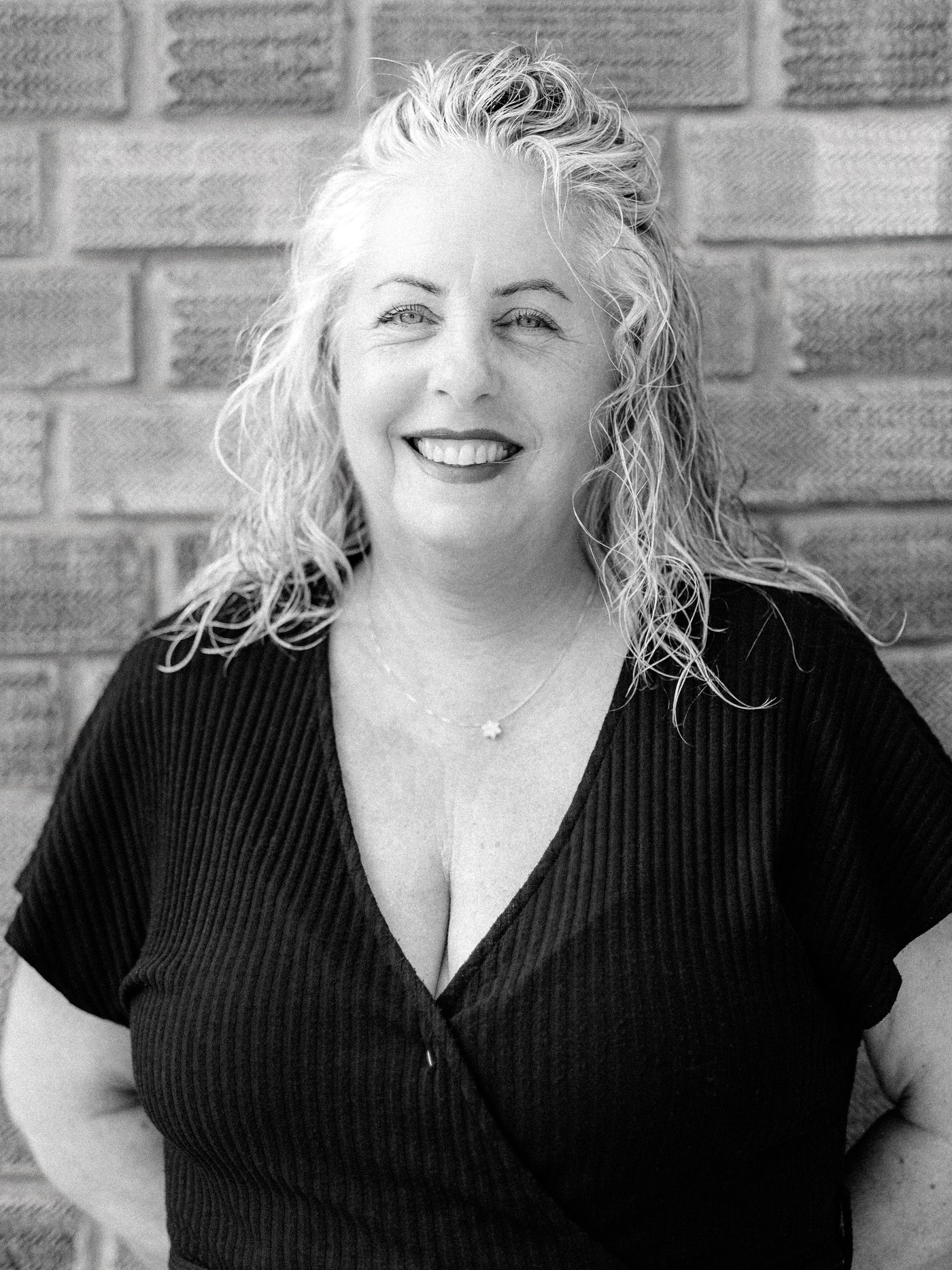 A black and white portrait of Method Salon senior stylist, Laura, a smiling woman with curly hair, wearing a dark ribbed wrap top, standing against a brick wall.