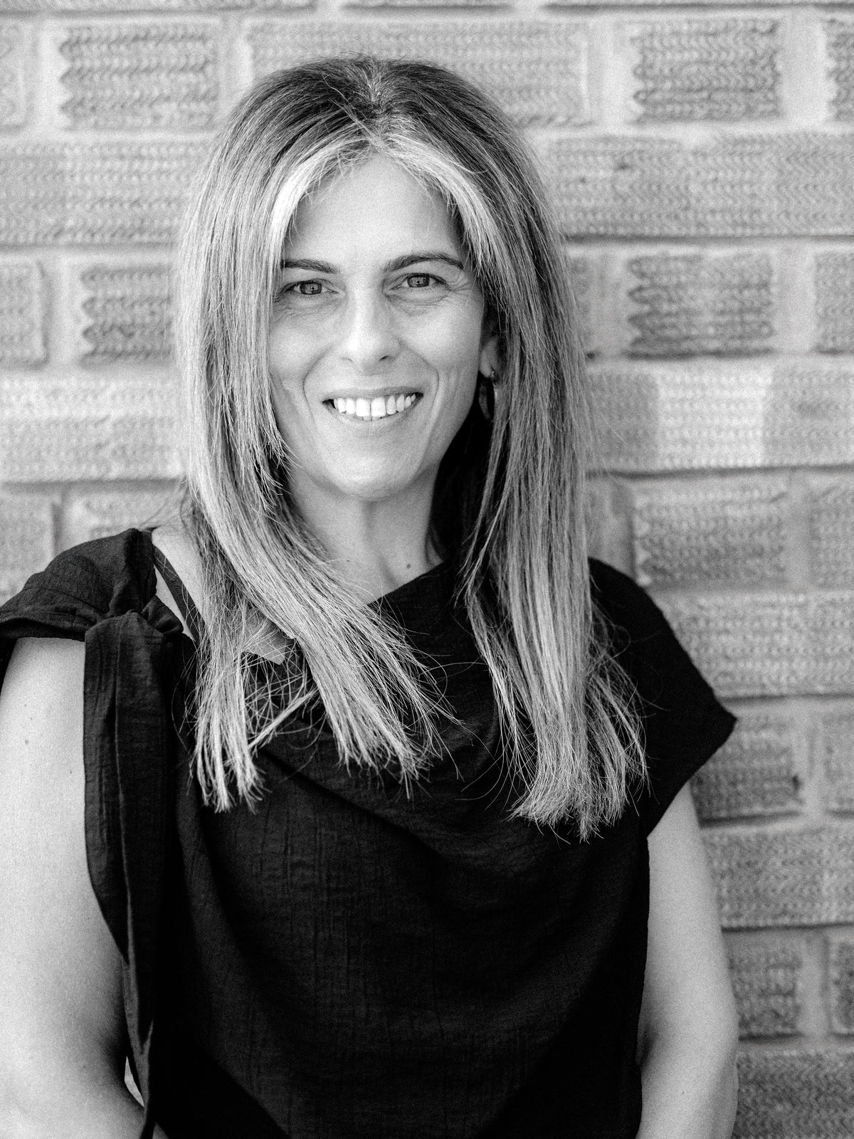 A black and white portrait of Method Salon senior stylist and colourist Lyndsey, a smiling woman wearing a black top, posing in front of a brick wall.