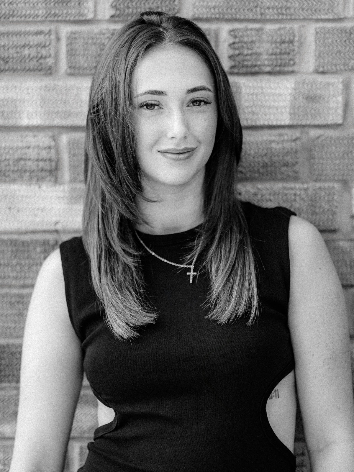 A black and white portrait of Method stylist and colourist Millie, a woman with shoulder-length hair wearing a sleeveless black top and a cross necklace, standing against a brick wall.