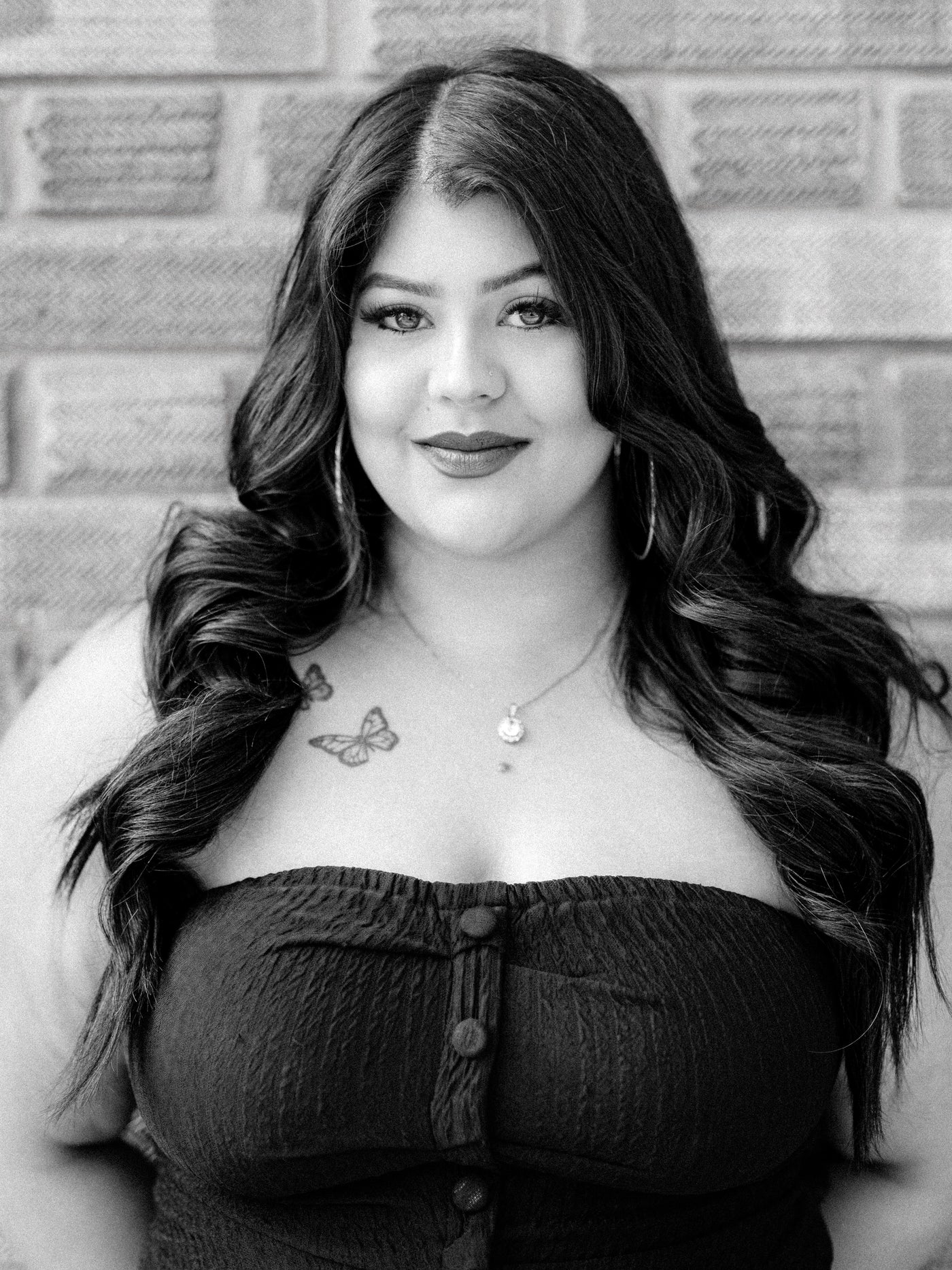 A black and white portrait of Method Salon senior stylist, Poonam, a woman with long wavy hair, wearing a strapless button-front top, standing in front of a brick wall.