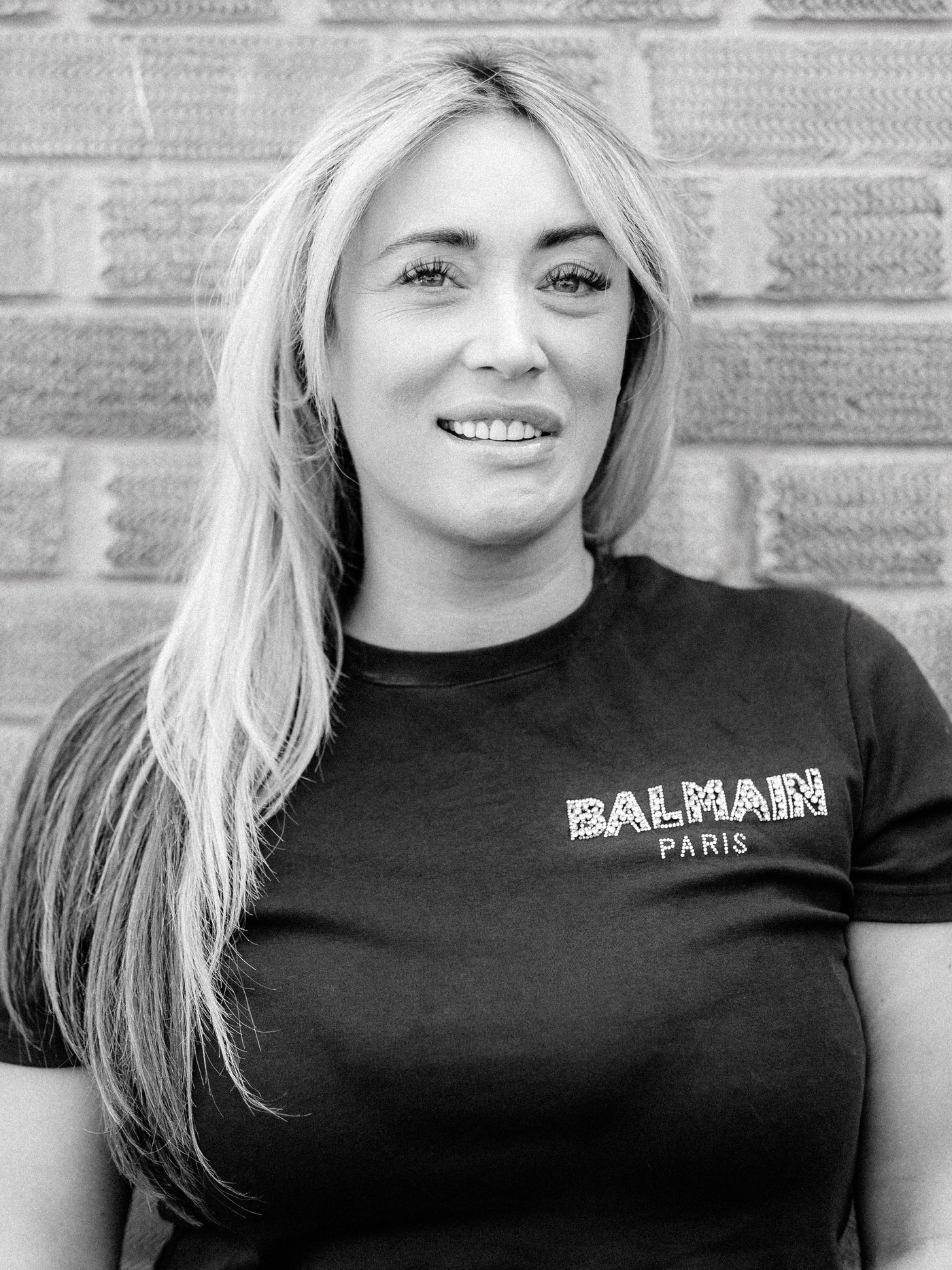 Method colourist Sasha, a woman with long blonde hair smiling in front of a brick wall, wearing a black Balmain Paris t-shirt. The image is in black and white.