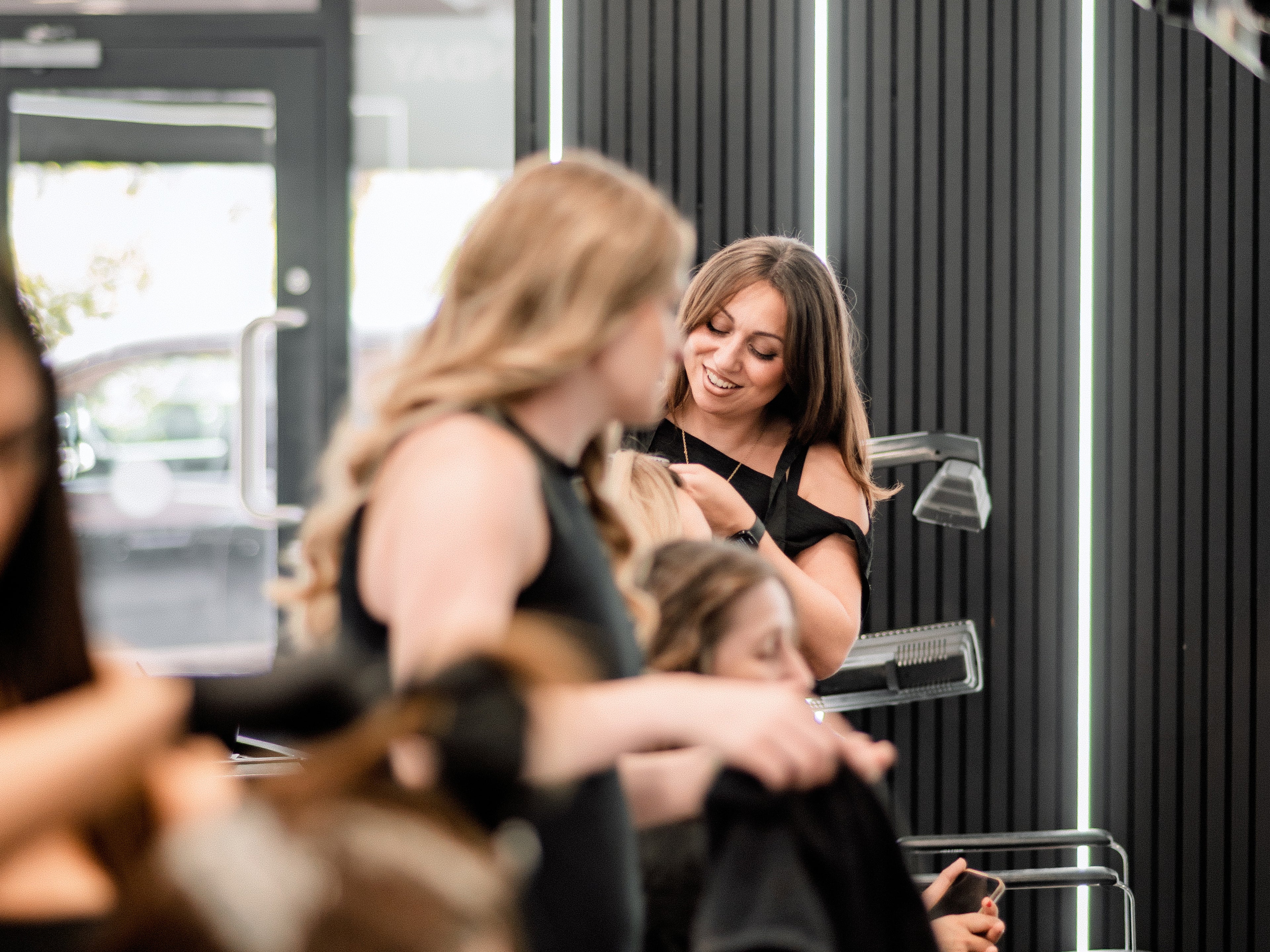 A salon interior scene with multiple people visible, including a stylist working on a client's hair in front of a dark slatted wall with vertical light strips.