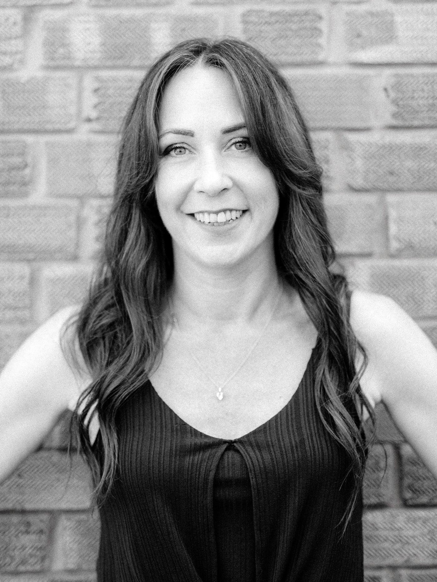 A black and white portrait of Method senior stylist Kelly, a smiling woman with long wavy hair, wearing a dark ribbed tank top, standing in front of a brick wall.