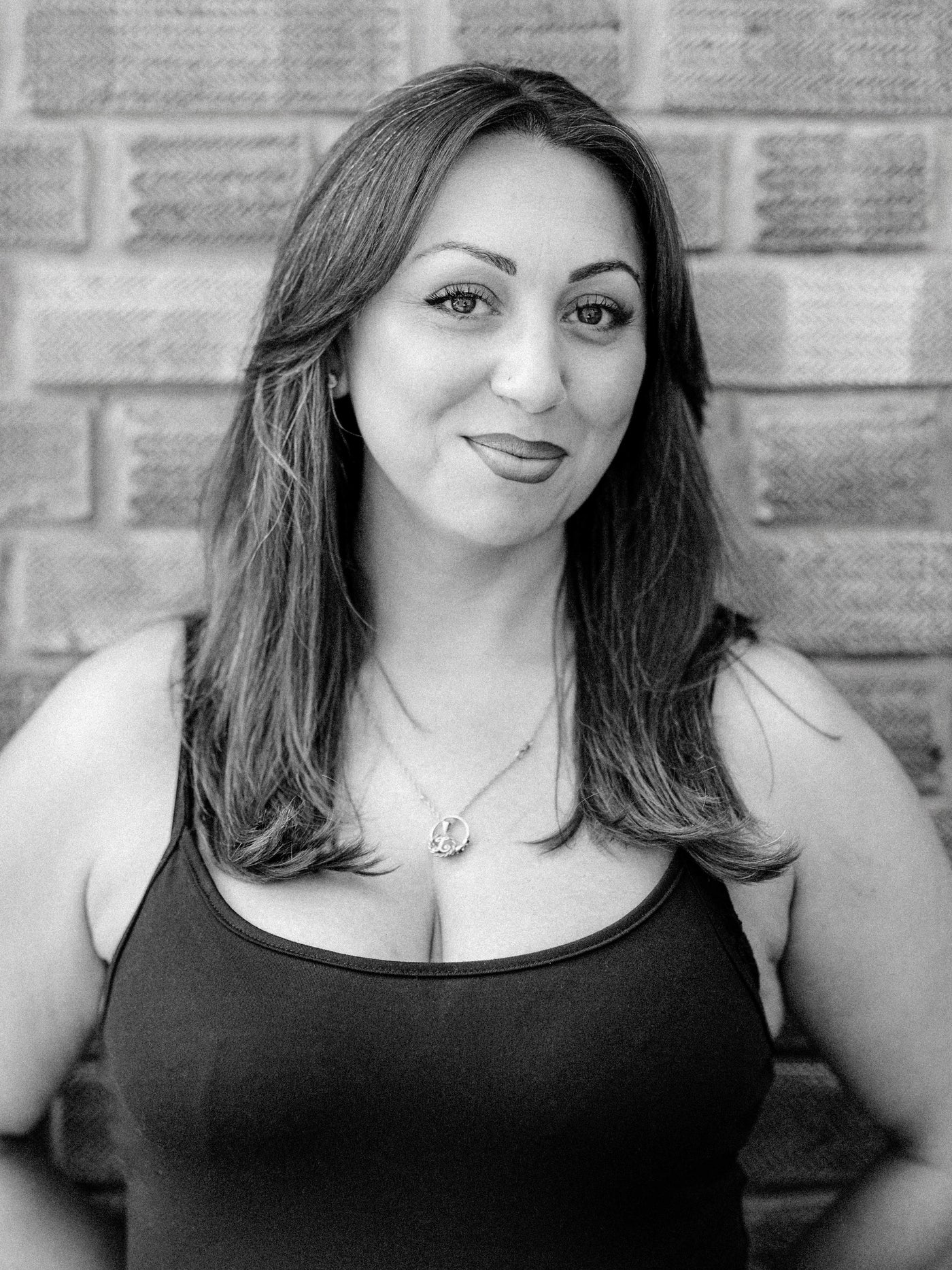 A black and white portrait of Method stylist and colourist Leah, a woman with shoulder-length hair wearing a black vest top and a necklace, standing against a brick wall.