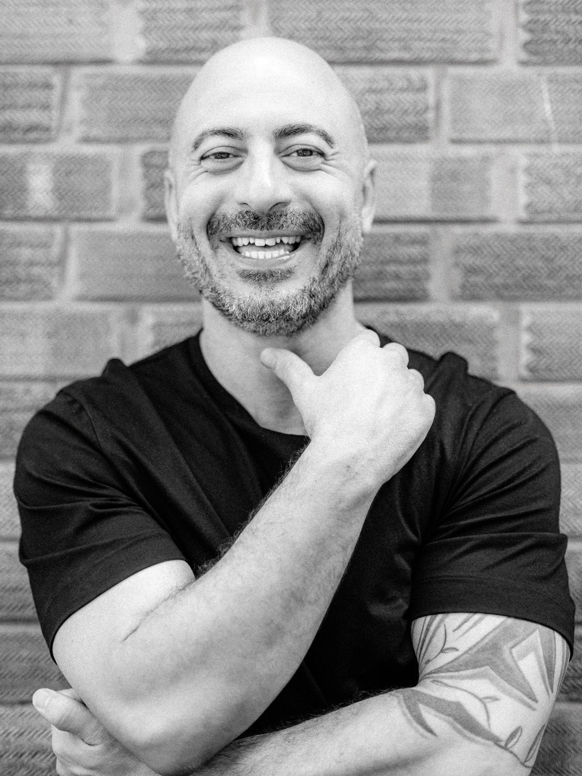 A black and white portrait of Anthony, Method salon director, a smiling man with a beard, wearing a black t-shirt, posing in front of a brick wall.