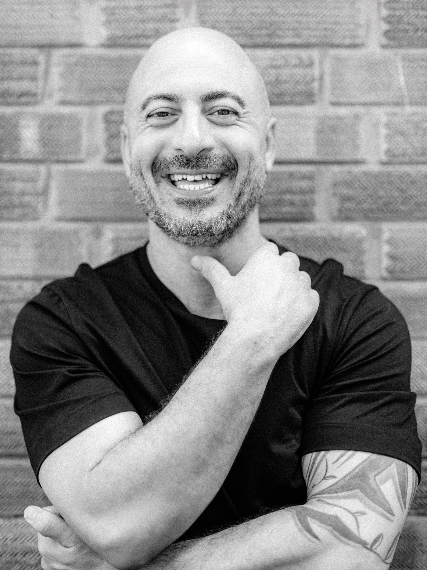 A black and white portrait of Anthony, Method salon director, a smiling man with a beard, wearing a black t-shirt, posing in front of a brick wall.